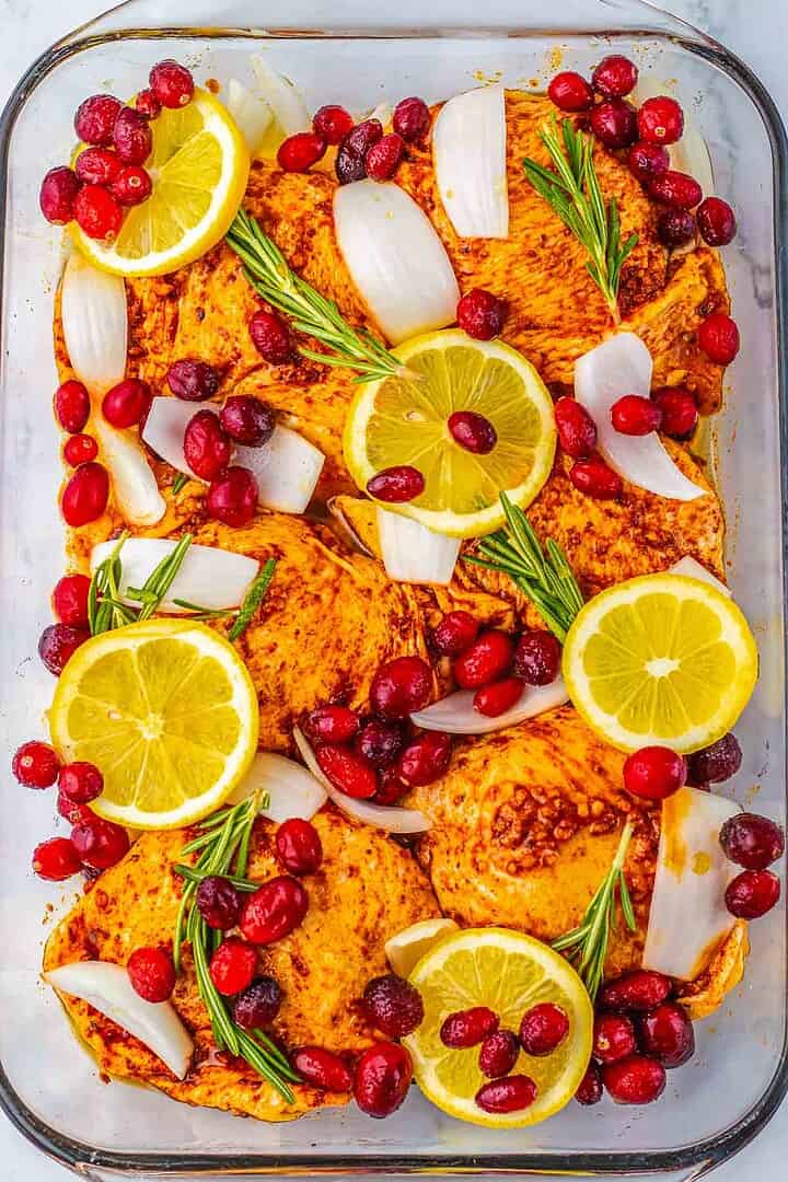 Raw chicken thighs in a dish with lemon slices, cranberries, onion chunks, and rosemary sprigs—perfect for making Roasted Cranberry Chicken bursting with flavor.
