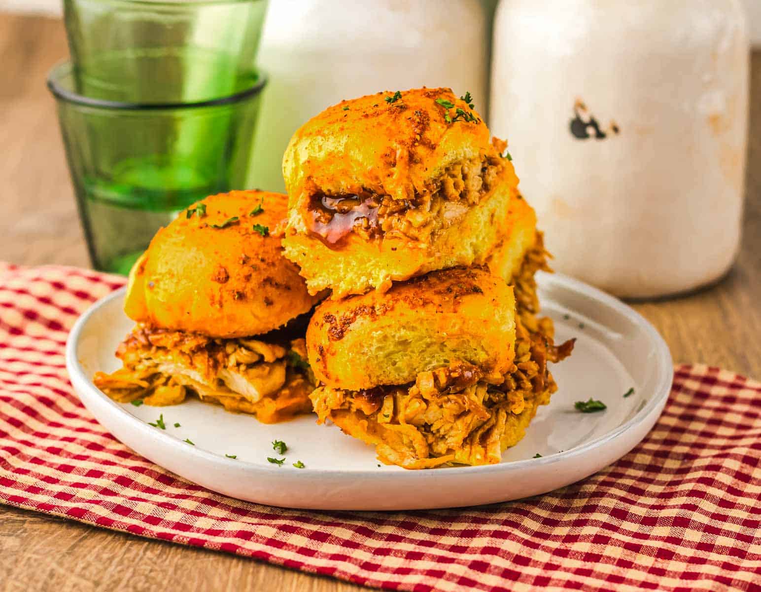 Three Cranberry BBQ turkey sliders on a plate, set on a red checkered cloth with green glasses in the background—a perfect slow cooker treat for any gathering.