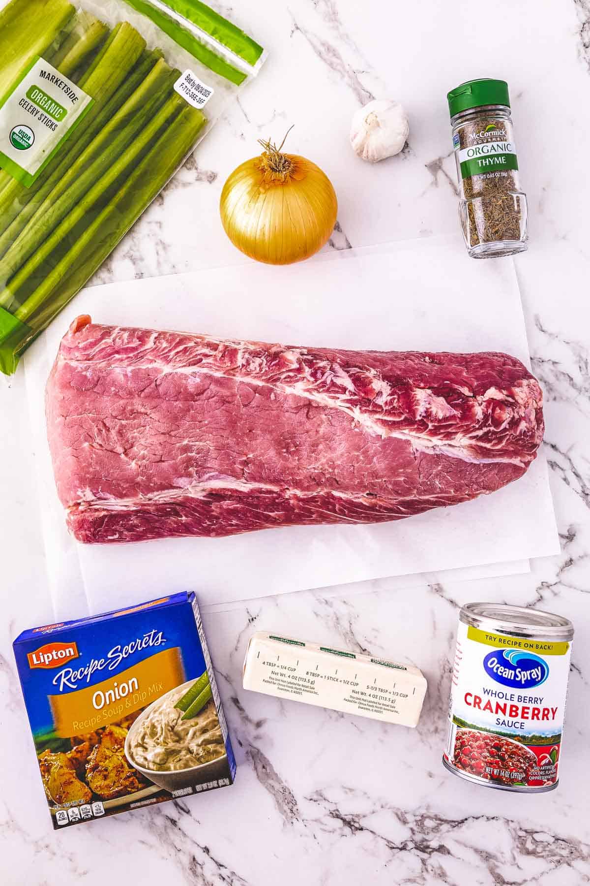 Raw pork loin, onion, celery, butter, cranberry sauce, onion soup mix, dried thyme, and garlic are ready to create a flavorful Cranberry Pork Loin perfect for your Crockpot on a marble surface.