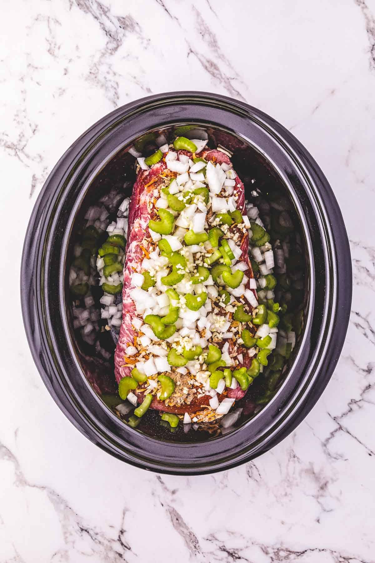 Raw pork loin topped with chopped onions and green peppers in a crockpot on a marble countertop.
