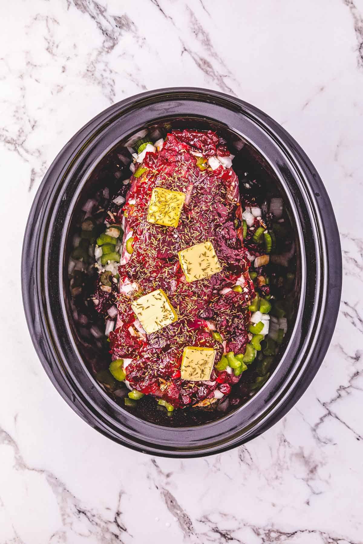 Raw meatloaf with herbs, ketchup, butter, and vegetables in a Crockpot on a marble countertop.