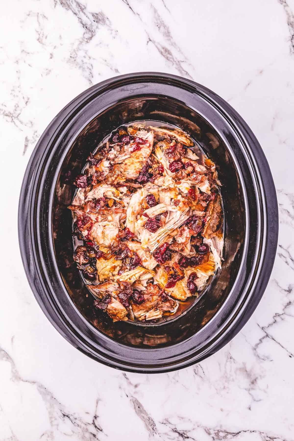 Shredded turkey with cranberries and juices in a black Crockpot on a marble countertop.