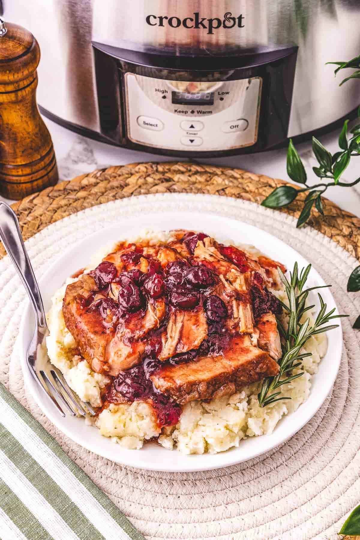 A plate of mashed potatoes topped with tender Crockpot pork loin and cranberry sauce, garnished with a sprig of rosemary nearby.