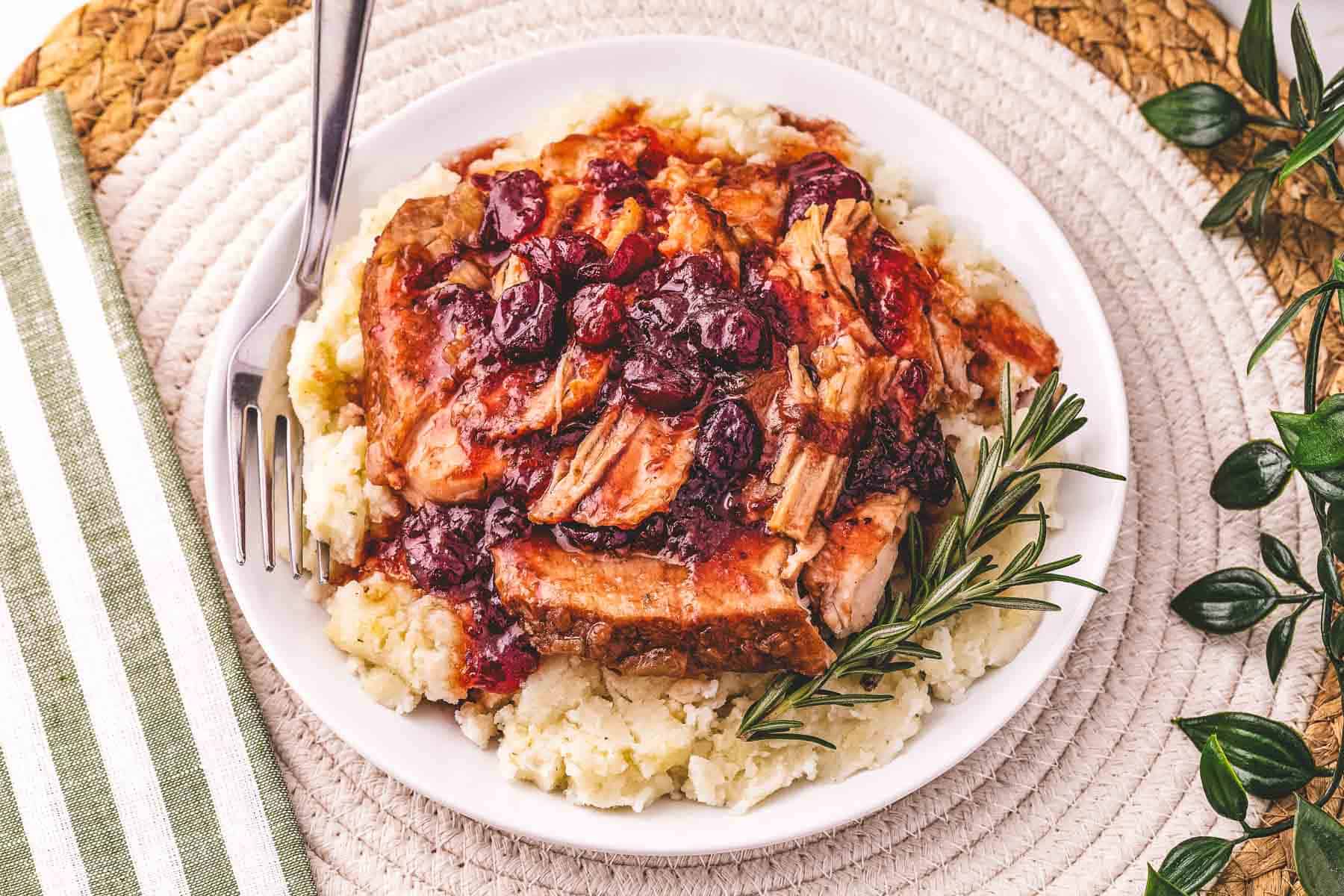 Plate of tender Crockpot pork loin with cranberry sauce over mashed potatoes, garnished with rosemary, and a fork on the side.
