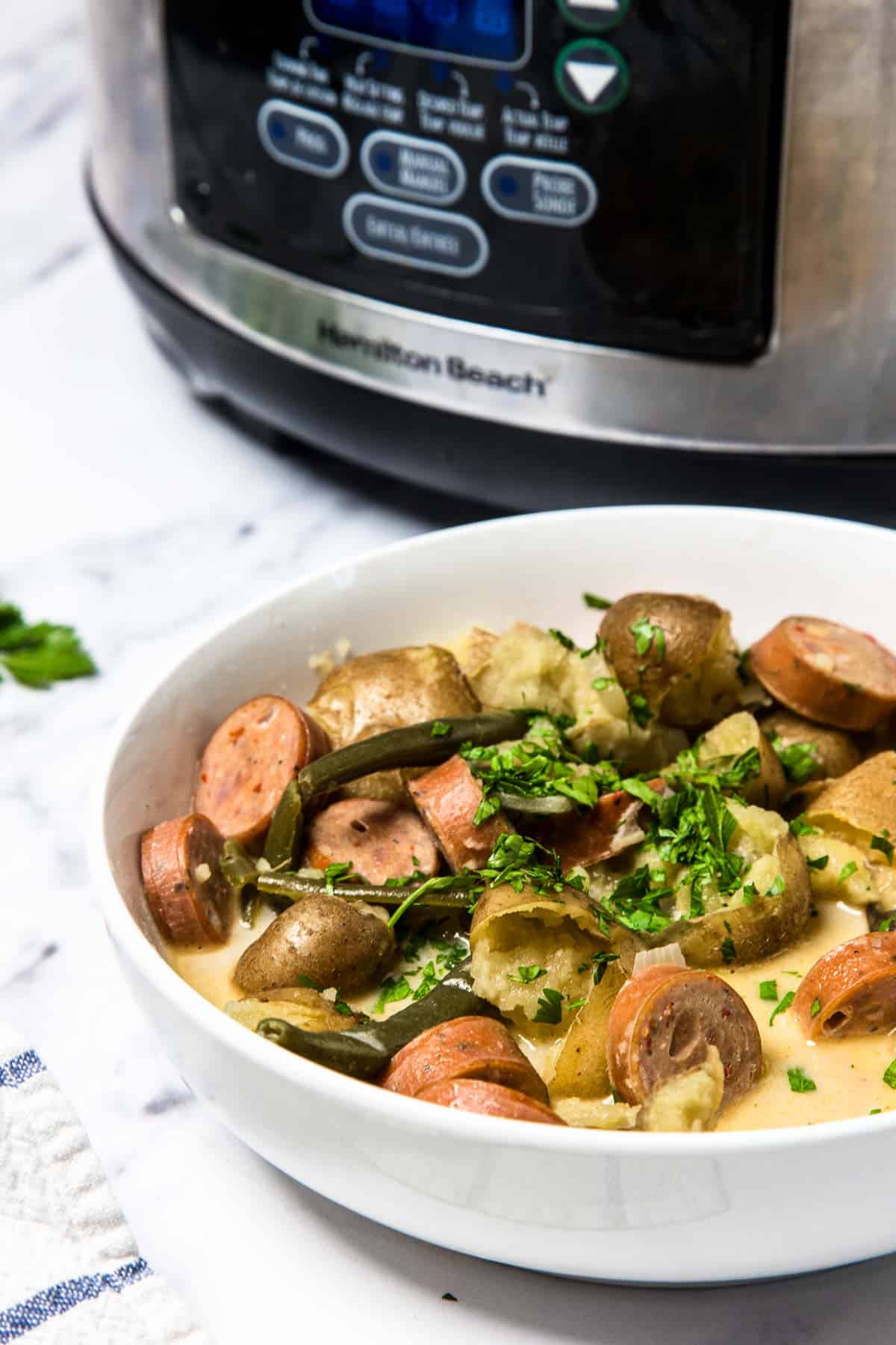 A bowl of creamy smoked sausage and potato soup, garnished with herbs, sits invitingly before a slow cooker in the background&mdash;perfect for savoring cozy flavors of swamp potatoes.