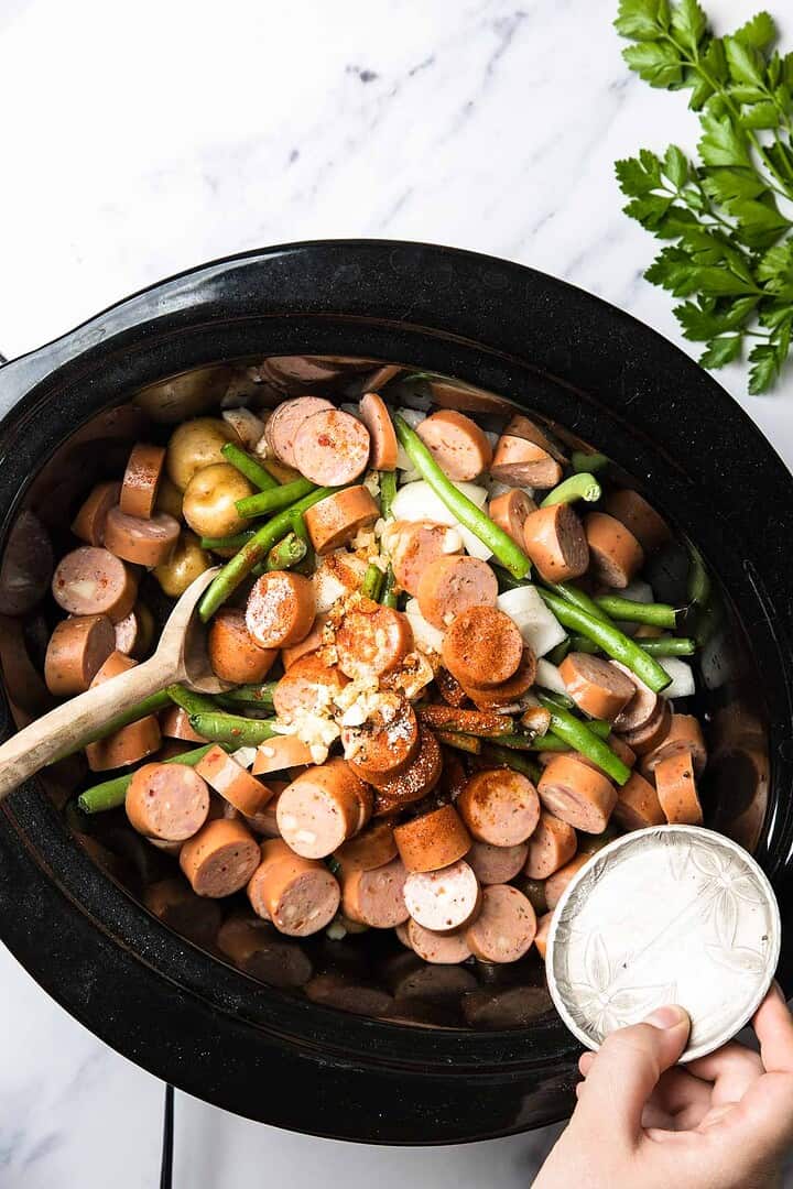 Smoked sausage, green beans, and swamp potatoes in a slow cooker being seasoned, with a wooden spoon and fresh herbs nearby.
