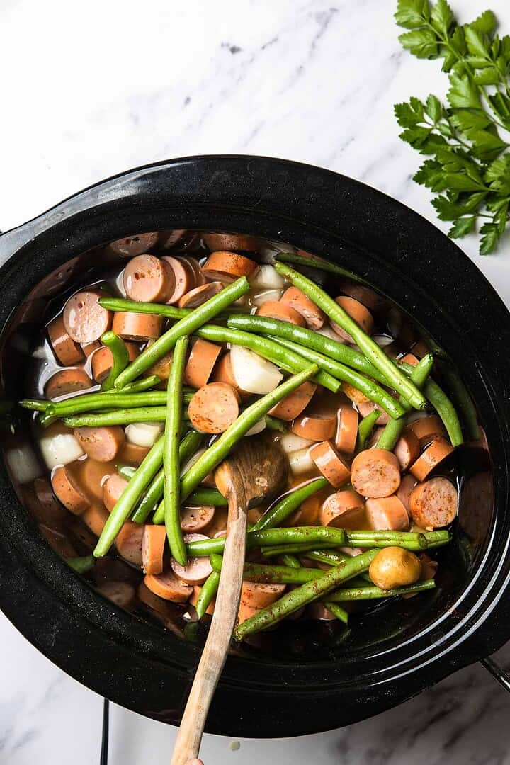 Sliced smoked sausage, green beans, and slow cooker potatoes simmer together in a cozy dish, stirred gently with a wooden spoon.