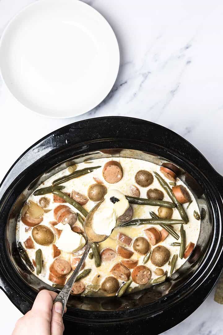 A hand holds a spoon with sour cream over Slow Cooker Swamp Potatoes&mdash;a hearty blend of slow cooker potatoes, smoked sausage, and green beans in a creamy sauce.