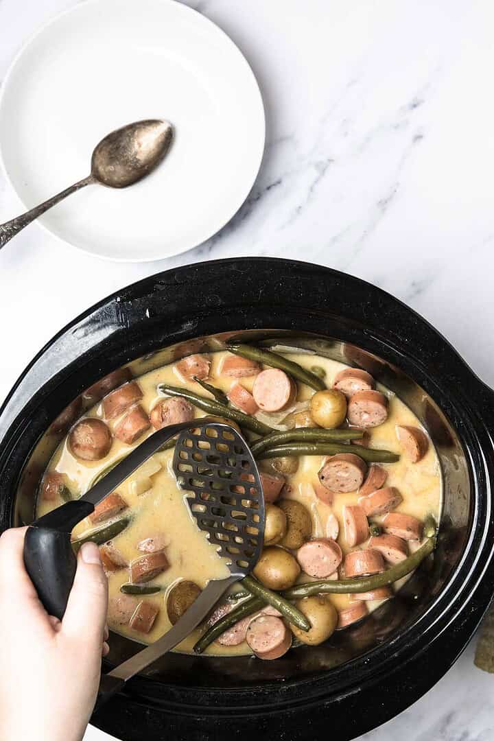 A hand mashes swamp potatoes in a slow cooker with smoked sausage, green beans, and creamy sauce.
