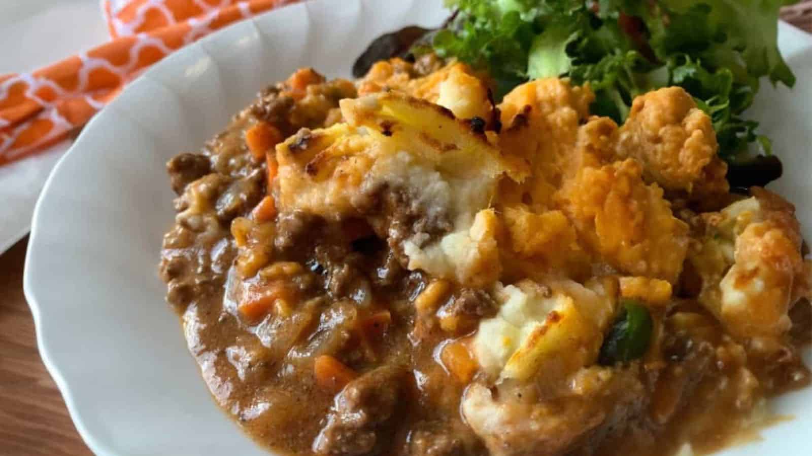 A plate of shepherd's pie with ground meat, vegetables, mashed potatoes, and a side of green salad.