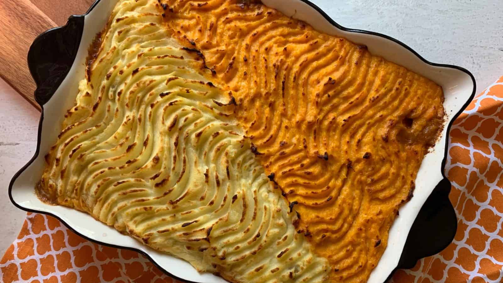 A plate with cottage pie casserole and a side of mixed greens on a white surface, accompanied by an orange-patterned cloth in the background.
