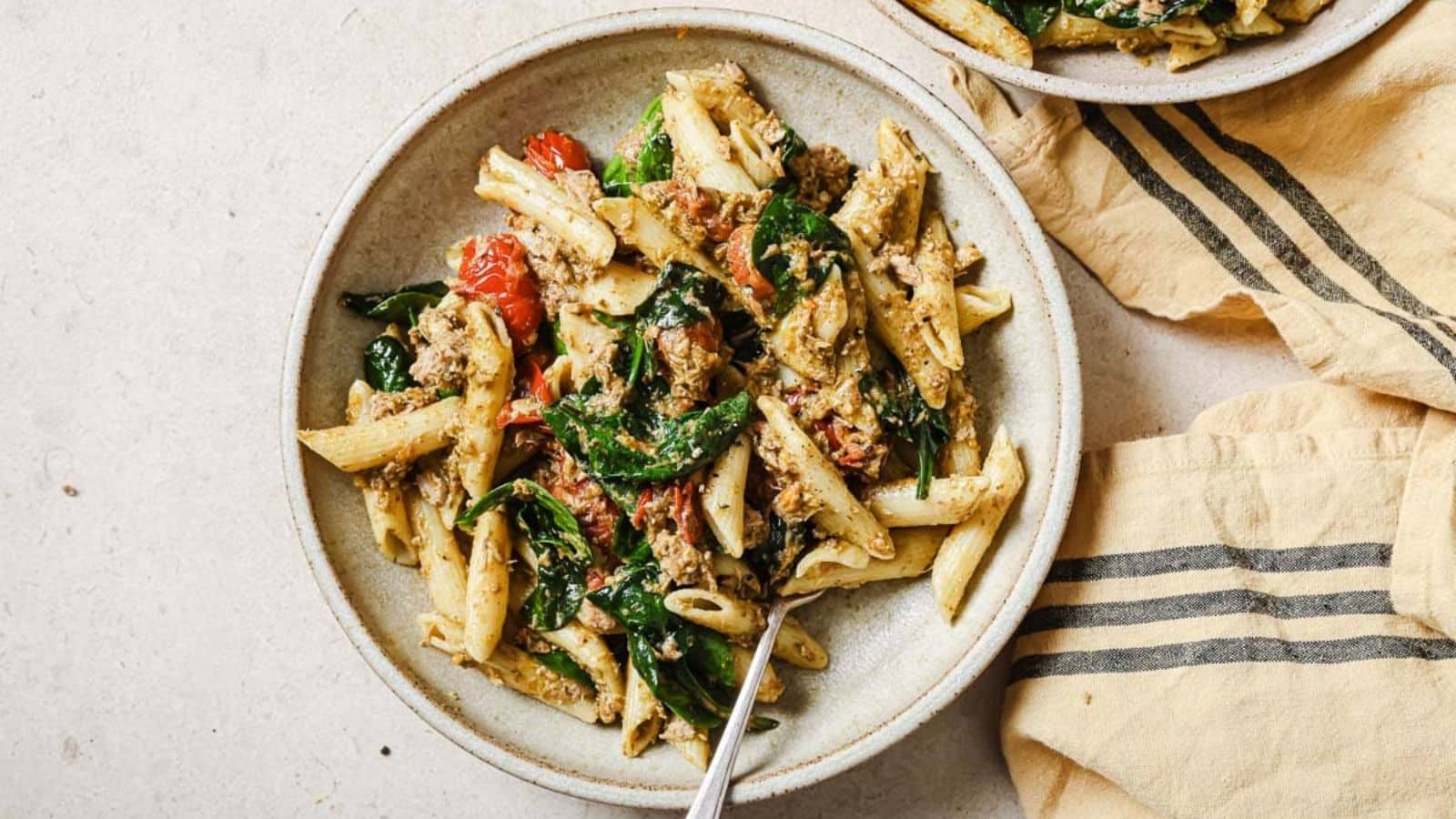 A plate of tuna pesto pasta with cherry tomatoes, a copper fork, and a pink linen next to a pan full of tuna pesto pasta.
