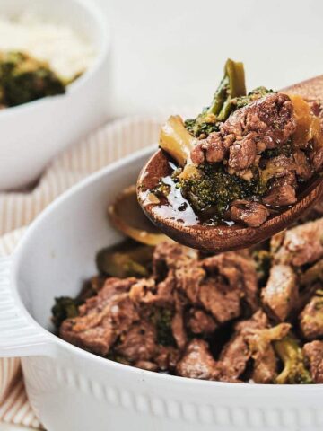 A wooden spoon holds beef and broccoli over a white casserole dish with more of the dish in the background.