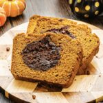 Two slices of Pumpkin Banana Bread with a chocolate swirl rest on a wooden cutting board, pumpkins in the background adding a festive Halloween touch.