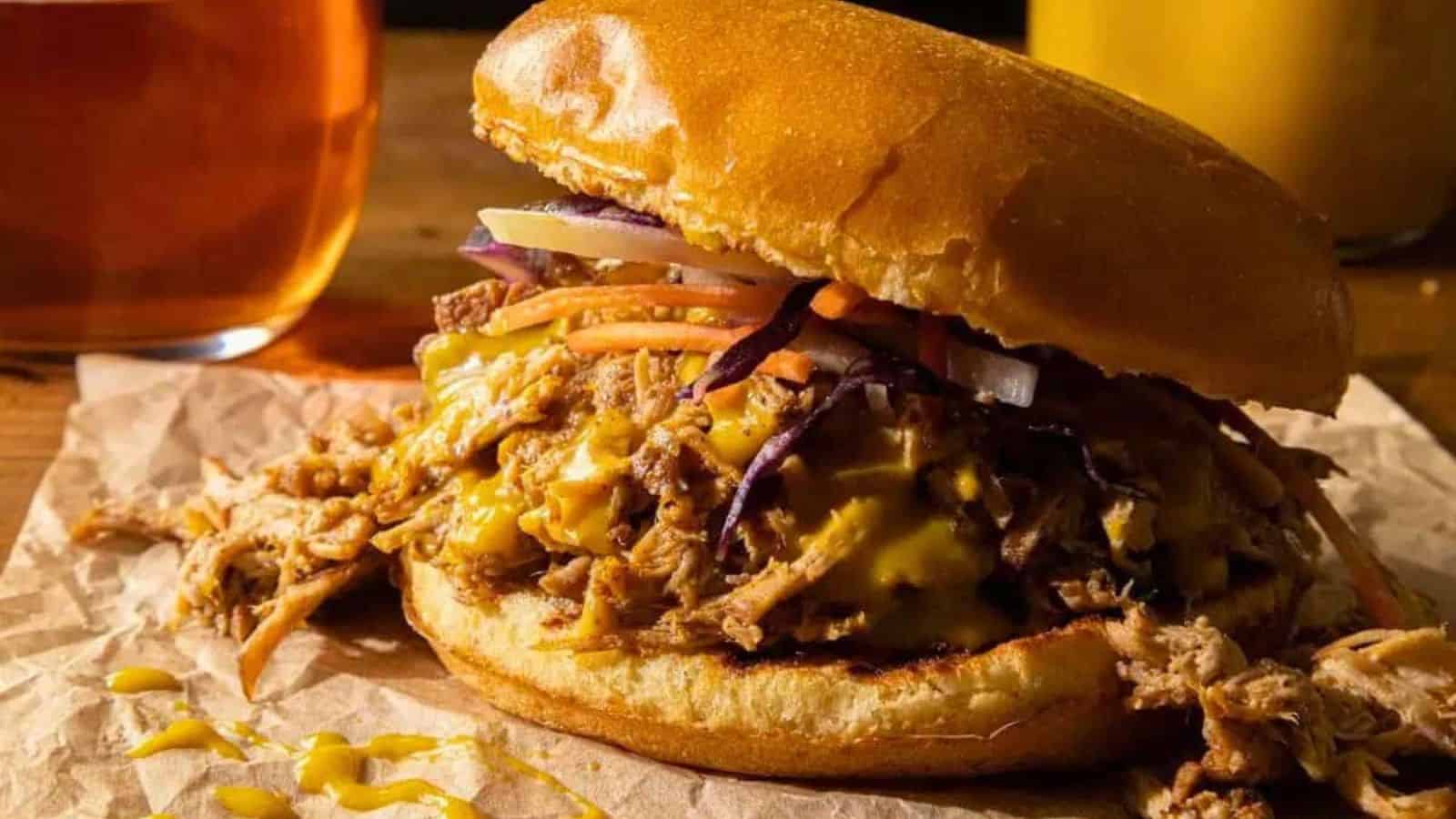 A delicious showcase of Southern cooking, this pulled pork sandwich with melted cheese, pickles, and shredded veggies is served on a bun with brown paper&mdash;perfect alongside a glass of beer and a yellow drink in the background.