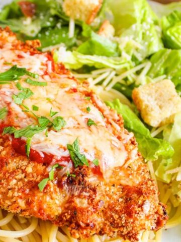 Plate of chicken parmesan on spaghetti with Caesar salad and croutons on the side.