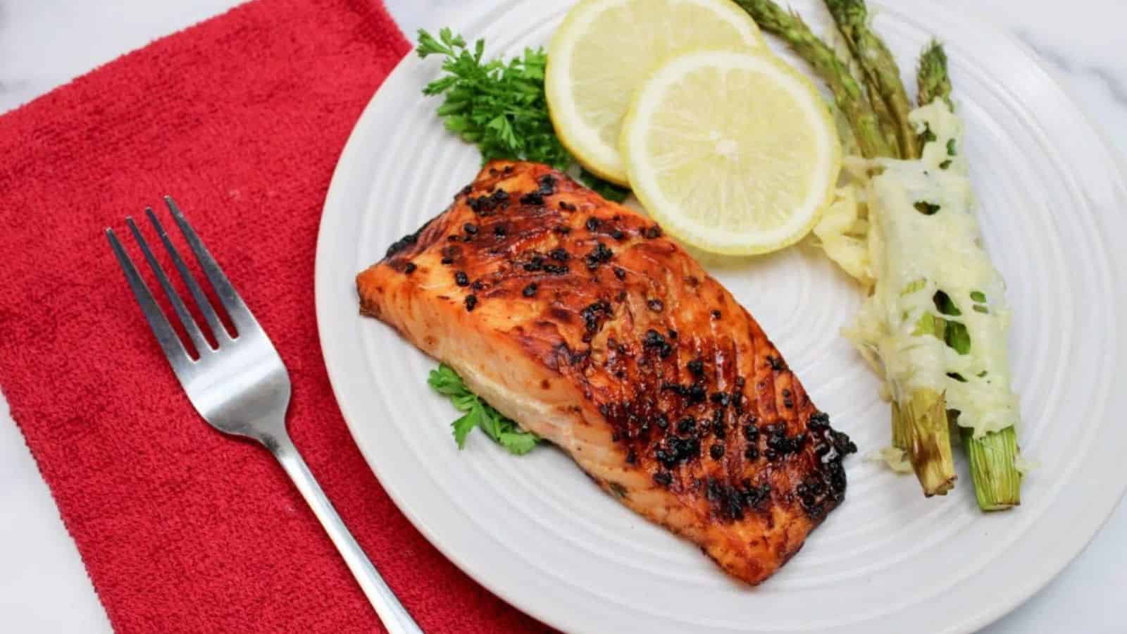 Grilled salmon with herbs, lemon slices, and asparagus on a white plate with a fork on a red napkin.
