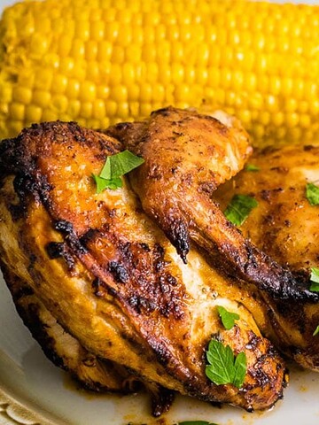 Grilled chicken with parsley, corn on the cob, and a bowl of orange sauce on a beige plate.