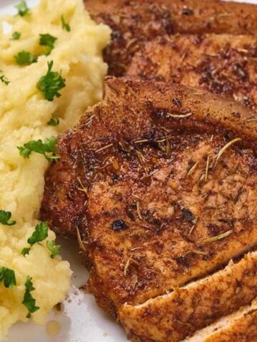 Two seasoned pork chops with mashed potatoes, garnished with parsley, on a white plate with a knife and fork.