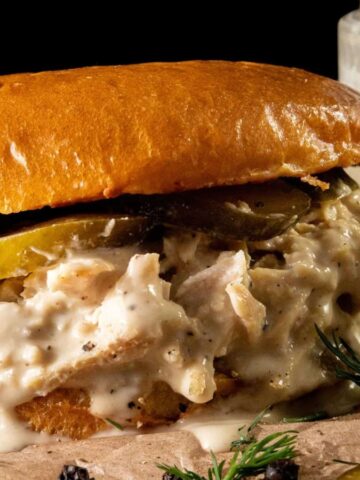 A creamy chicken sandwich with pickles on a bun, garnished with herbs, next to a jar of white sauce.