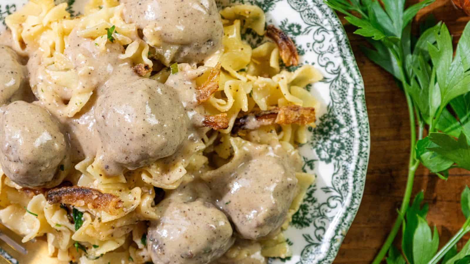 A close-up of a plate with egg noodles topped with easy Swedish meatballs and creamy gravy, garnished with herbs. Fresh parsley is visible in the background on a wooden surface.