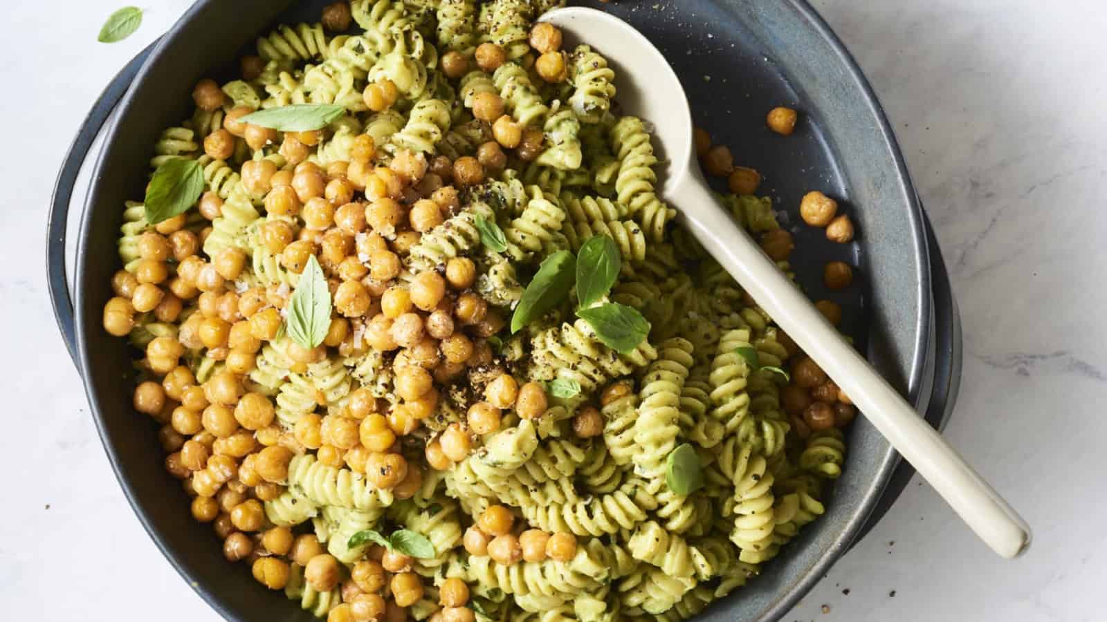A bowl of rotini pasta with green pesto sauce, crispy chickpeas, and fresh basil leaves, with a spoon.