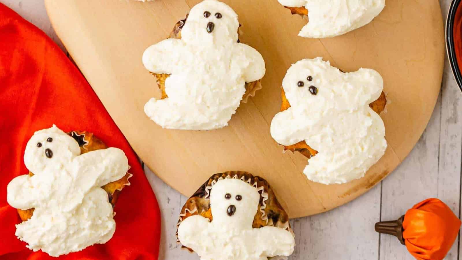 Cupcakes decorated with white frosting shaped like ghosts, displayed on a wooden board with a red napkin nearby.