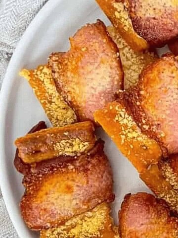A white plate of golden-brown bacon-wrapped crackers sprinkled with grated cheese.