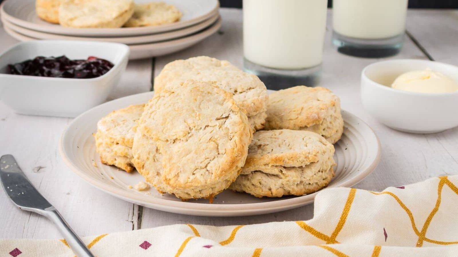 A plate of biscuits is set on a table with a knife, a bowl of butter, a bowl of jam, two glasses of milk, and a patterned napkin.