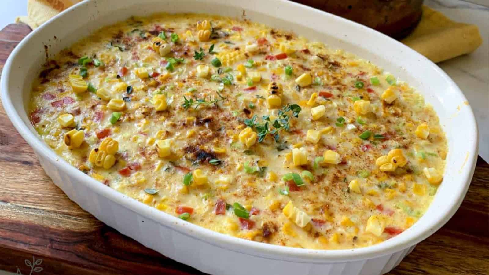 A baked corn casserole with chopped peppers and herbs is presented in a white oval dish on a wooden surface, with a patterned cloth nearby.