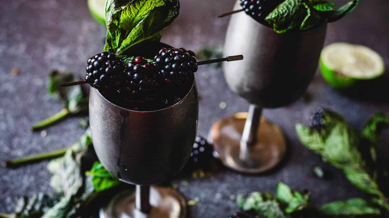 Two metal goblets filled with blackberries and mint, with scattered mint leaves around them.