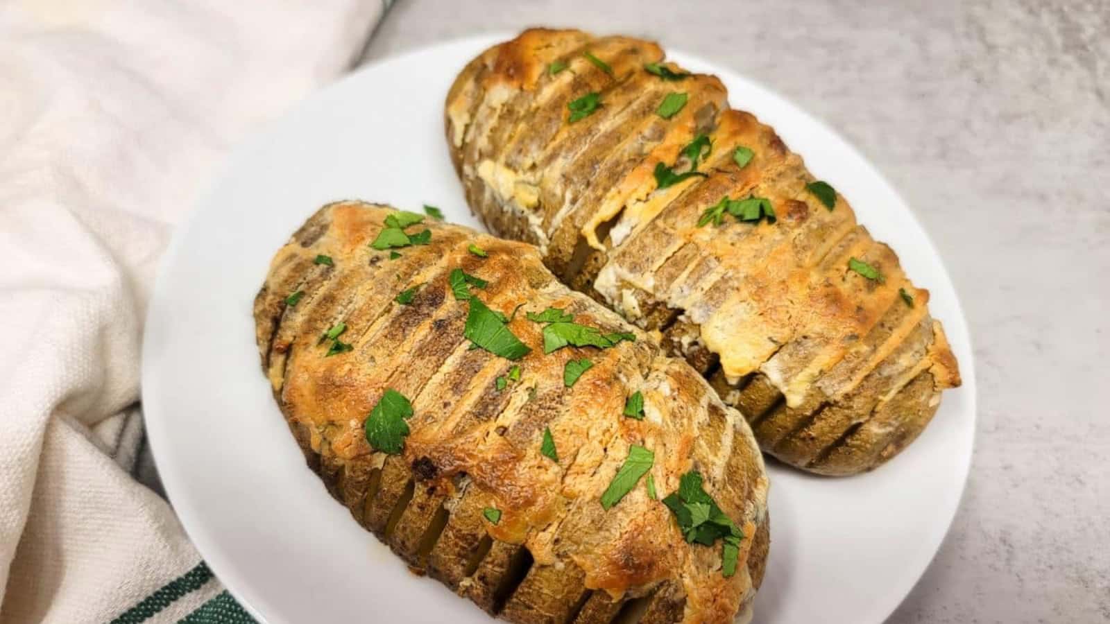 Two hasselback style potatoes topped with baked Boursin on a white plate with a hand holding them.