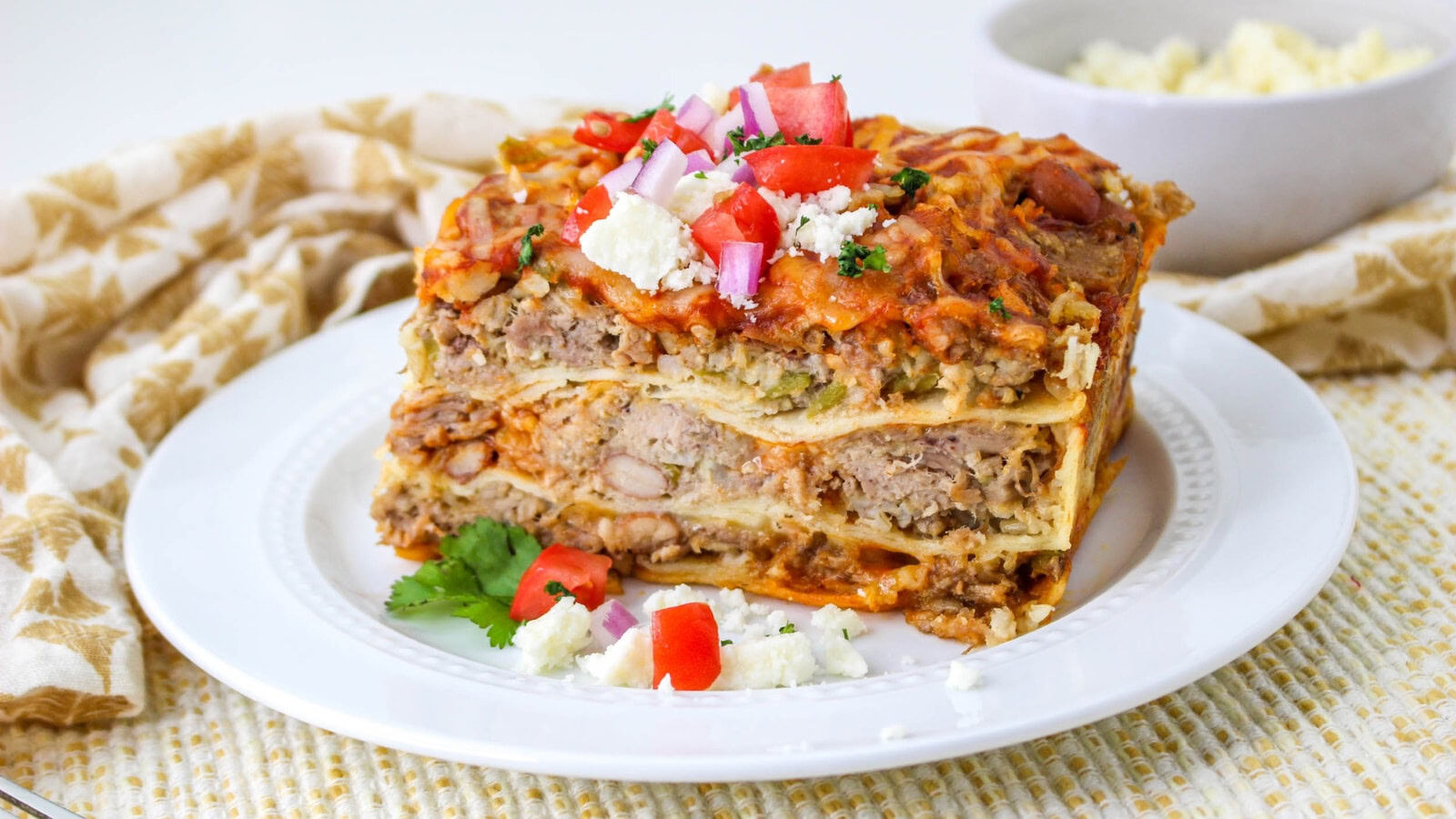 A slice of carnitas casserole on a white plate, topped with diced tomatoes, onions, and cheese. A bowl of cheese is in the background.