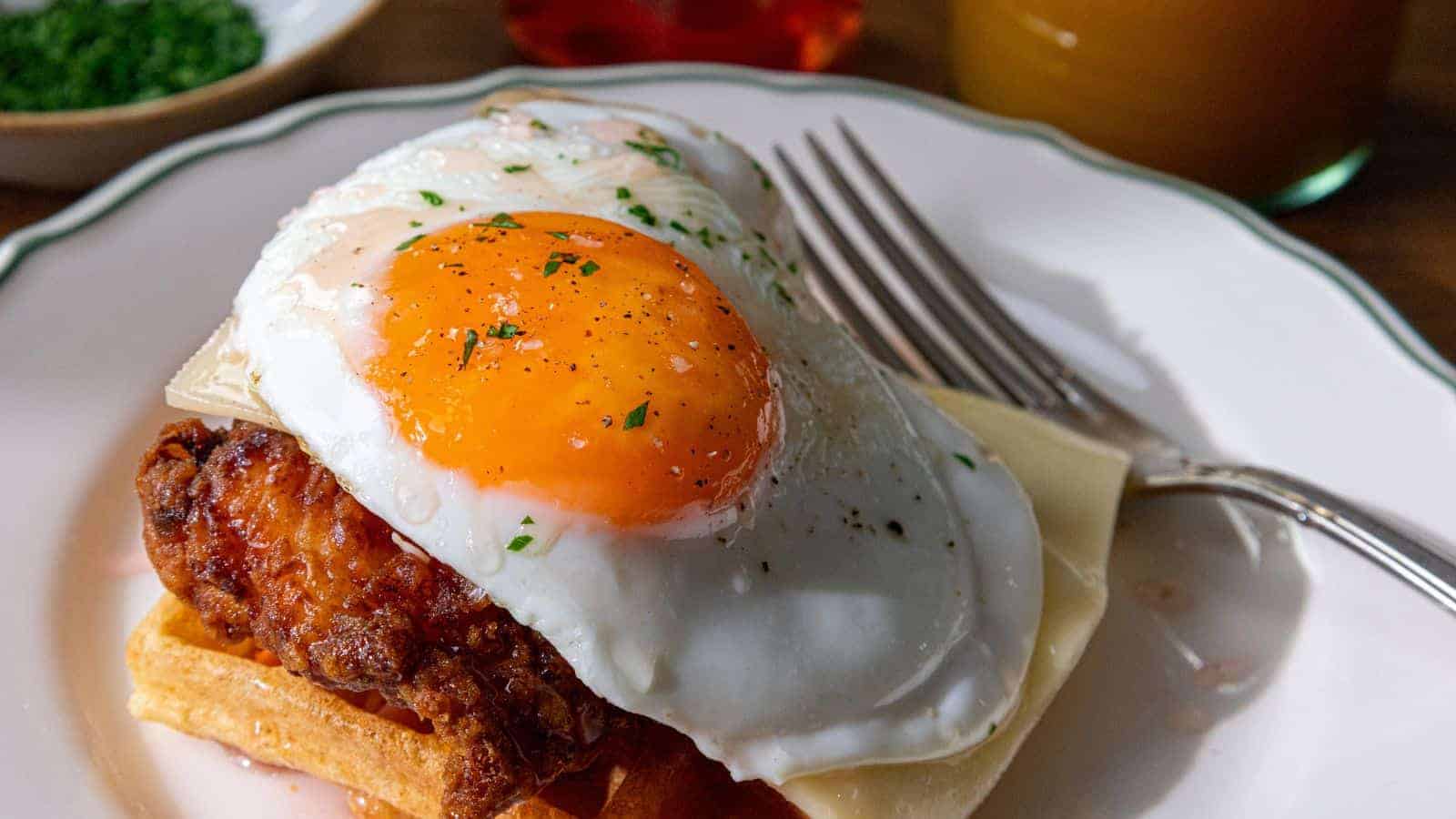 A close-up of a plate with a crispy waffle topped with fried chicken, melted cheese, and a sunny-side-up egg garnished with herbs. A fork rests on the plate's edge. In the background, there are blurred images of drinks and a small bowl.