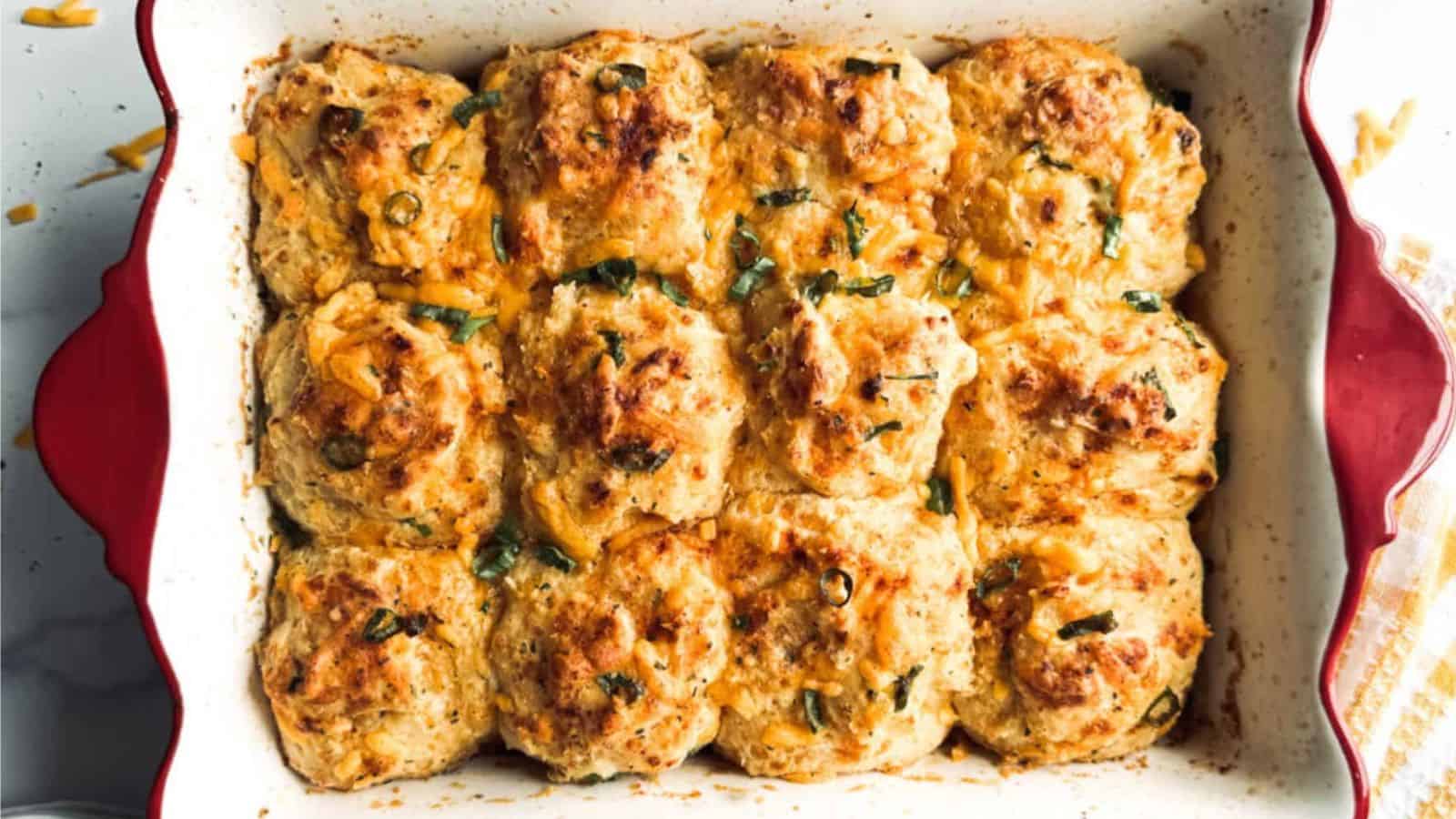 Golden-brown cheesy biscuits, sausage, and eggs topped with chopped herbs in a red floral-patterned baking dish.