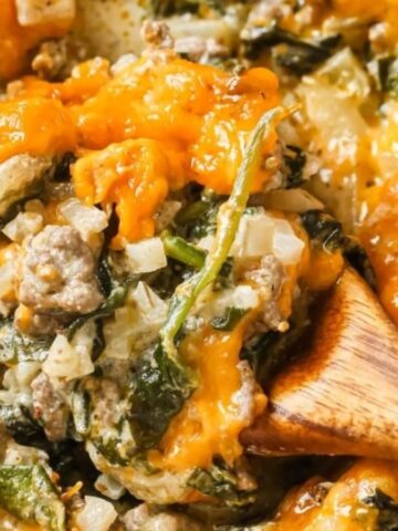 A wooden spoon scoops cheesy ground beef and spinach casserole with melted cheese on top.