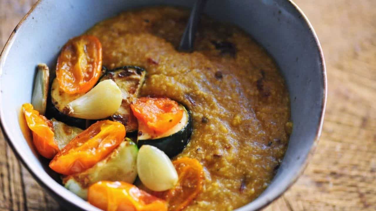 A bowl of thick soup with roasted tomatoes, zucchini slices, and garlic on top, with a spoon inside.