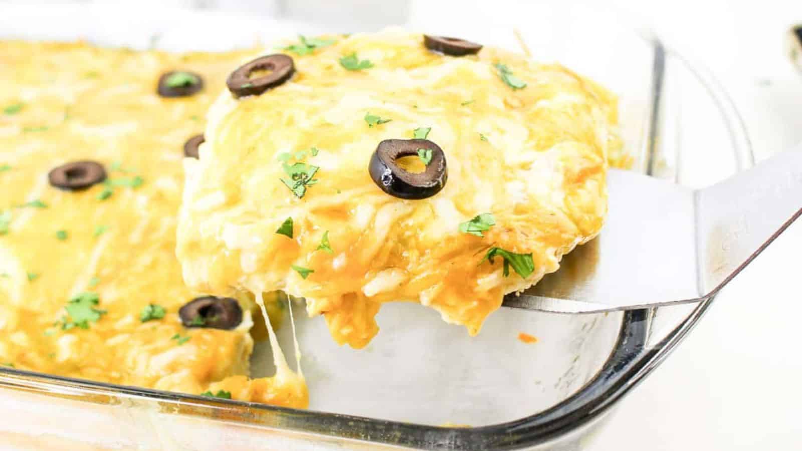 A slice of cheesy enchilada casserole with black olives and herbs is lifted from a glass baking dish.