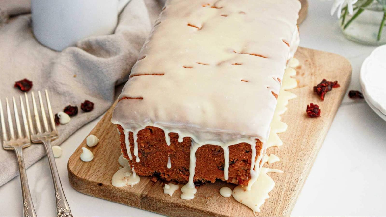 A loaf cake with white icing drips sits on a wooden board, with forks and dried berries nearby.