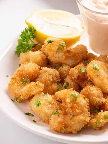 A plate of breaded fried shrimp with parsley, lemon, and dipping sauce in a glass cup.
