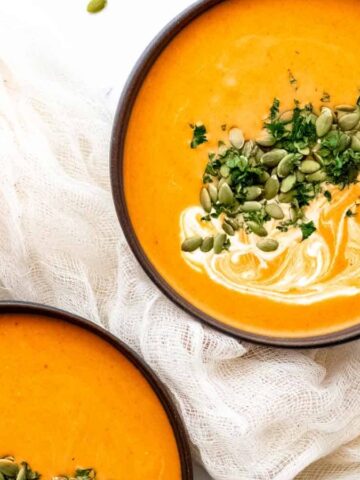 Two bowls of creamy pumpkin soup topped with seeds and herbs, next to a small pumpkin and a white cloth.