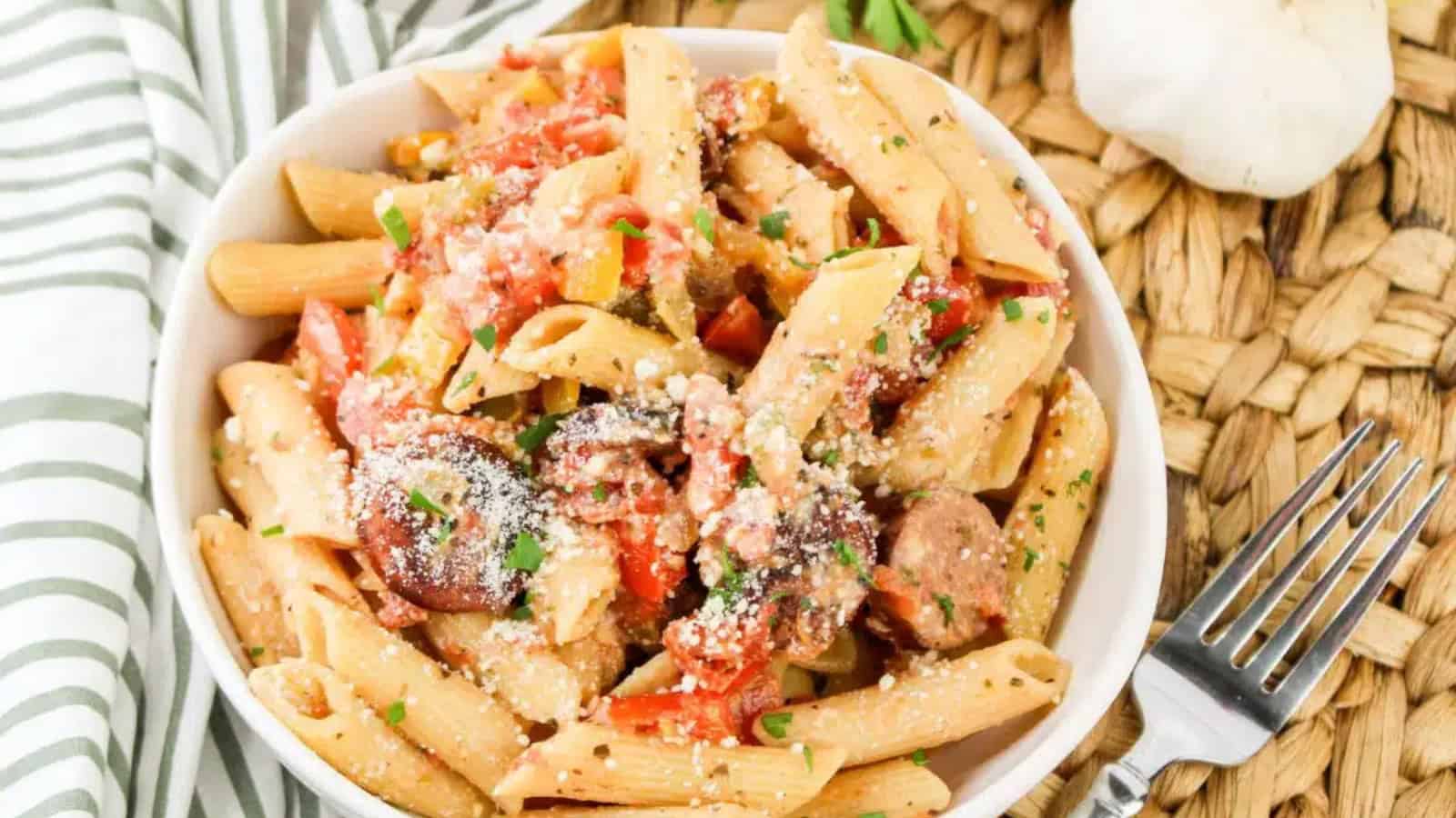 A bowl of penne pasta with sliced sausage, tomatoes, and grated cheese, garnished with herbs. A fork is placed beside the bowl.
