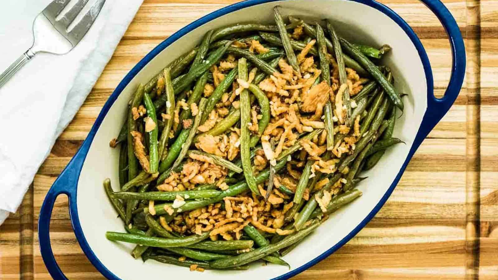 Oval dish with cooked green beans topped with crispy fried onions, on a wooden surface.