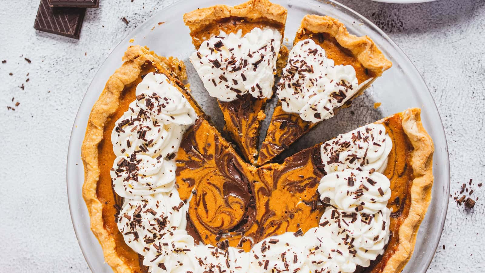 Pumpkin pie with chocolate swirl, topped with whipped cream and chocolate shavings, on a glass plate.