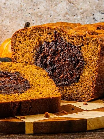 A sliced pumpkin bread loaf with a chocolate swirl center sits on a wooden board, surrounded by small pumpkins and autumn decorations.