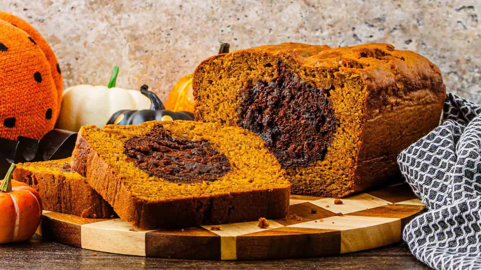 A sliced pumpkin bread loaf with a chocolate swirl center sits on a wooden board, surrounded by small pumpkins and autumn decorations.