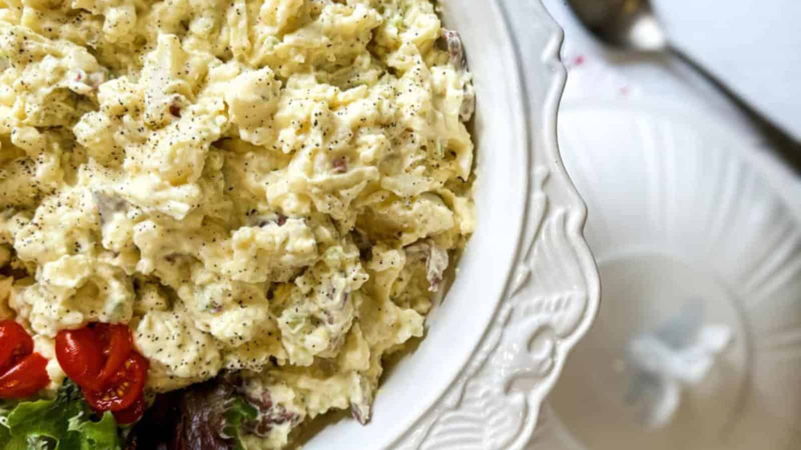 A bowl of creamy potato salad with specks of pepper, garnished with cherry tomatoes and lettuce on a decorative white plate.