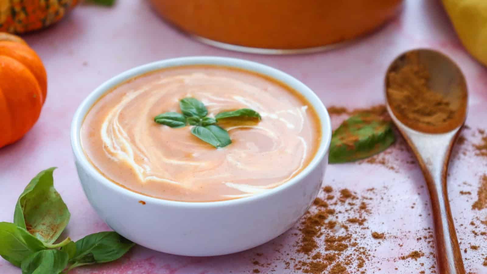 A ladle scoops creamy pumpkin soup from a glass pot, garnished with basil leaves.