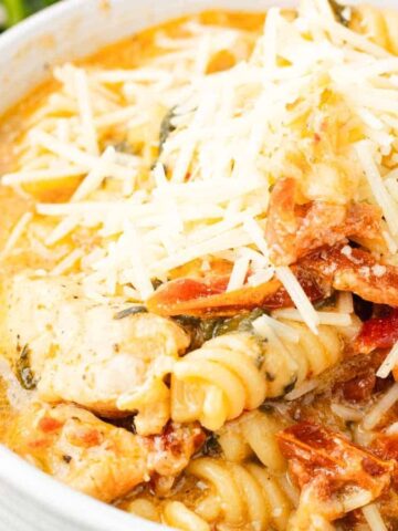 A bowl of creamy pasta with chicken, sun-dried tomatoes, spinach, and shredded cheese on top.