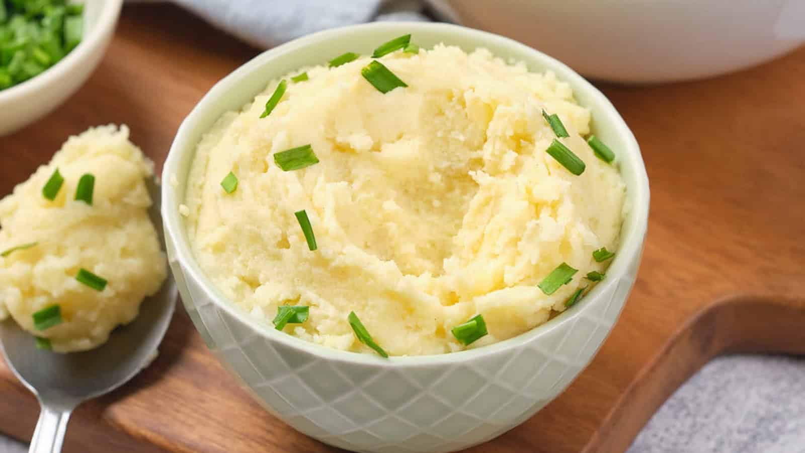 A bowl of mashed potatoes with chopped chives on a wooden board, accompanied by a spoon and a small dish of chives.